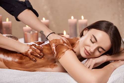 Application d’un soin au chocolat sur le dos d’une femme allongée dans une ambiance chaleureuse.