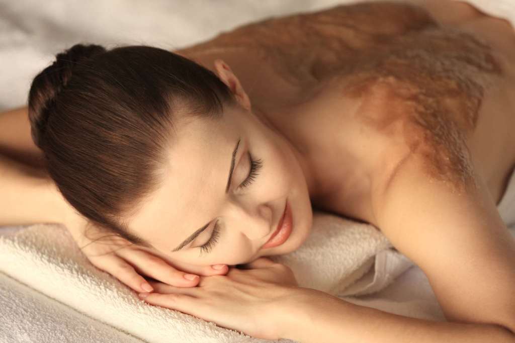 Soin exfoliant appliqué sur le dos d’une femme allongée dans une ambiance douce.
